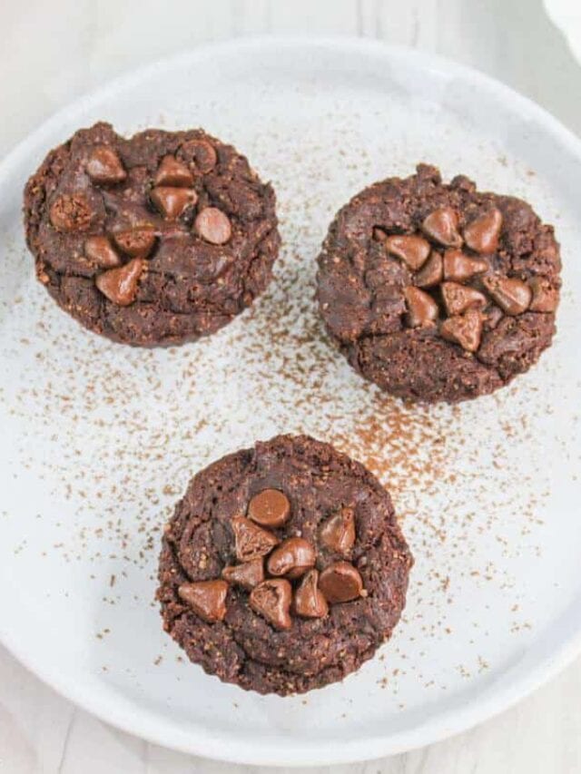 Three chocolate chip cookies on a white plate, dusted with cocoa powder, with visible chocolate chunks on top.
