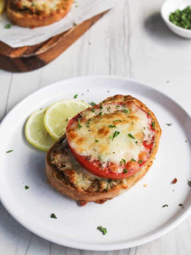 A baked tomato slice topped with melted cheese on a toasted bread, garnished with parsley, served on a white plate with a lime wedge.
