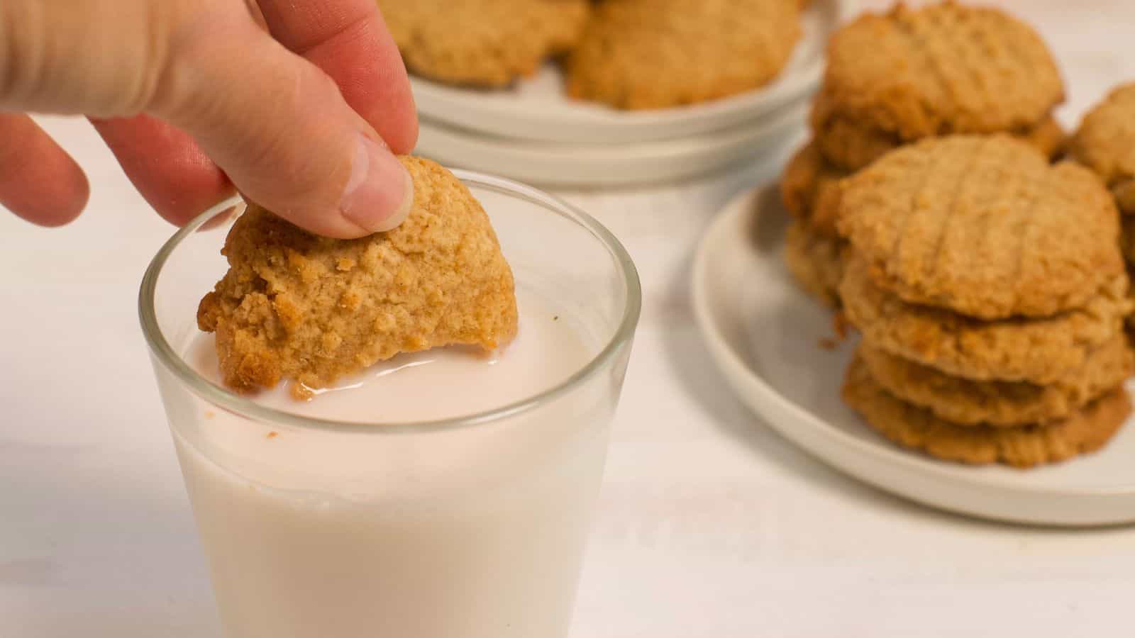 A hand dipping a peanut butter cookie into a glass of milk, with more cookies stacked on a plate in the background.