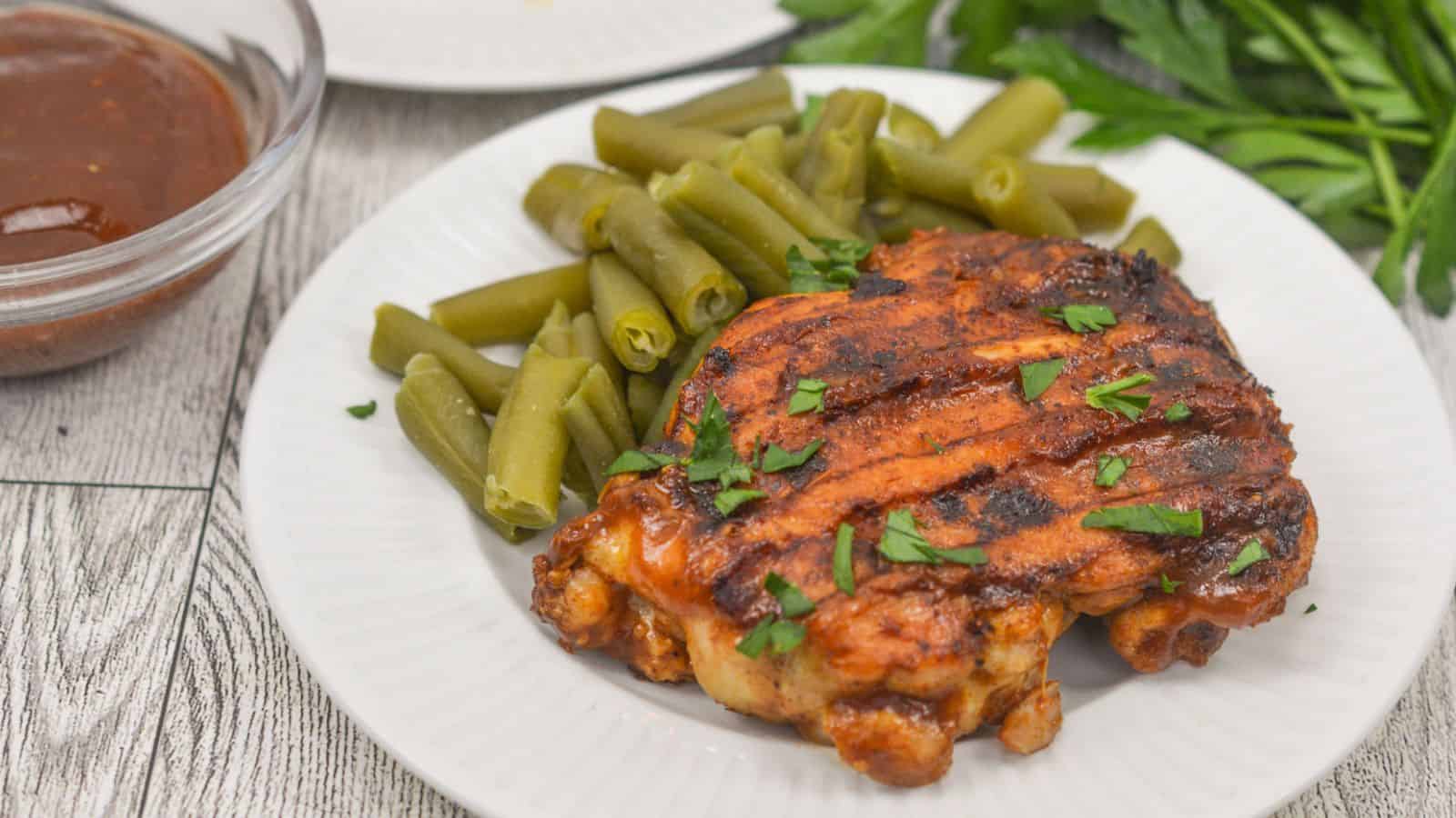 Grilled chicken thigh on a white plate with green beans and a side of barbecue sauce, garnished with chopped herbs on a wooden table.
