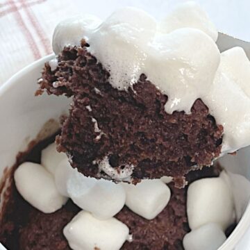 A spoon lifting a bite of chocolate mug cake topped with melted marshmallows from a white mug.