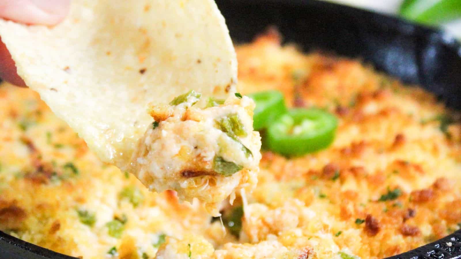 A close-up of a hand dipping a tortilla chip into a skillet of melted, cheesy jalapeño dip, with a golden crust and visible green jalapeño slices.