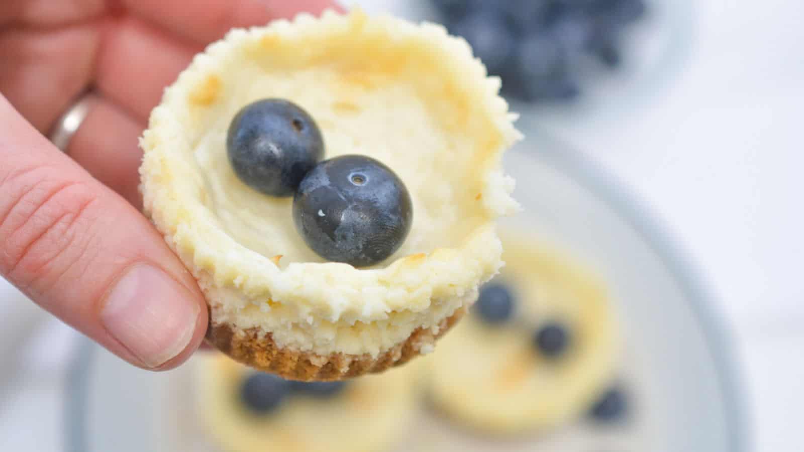 A hand holds a mini cheesecake topped with two blueberries, with more cheesecakes and berries on a plate in the background.