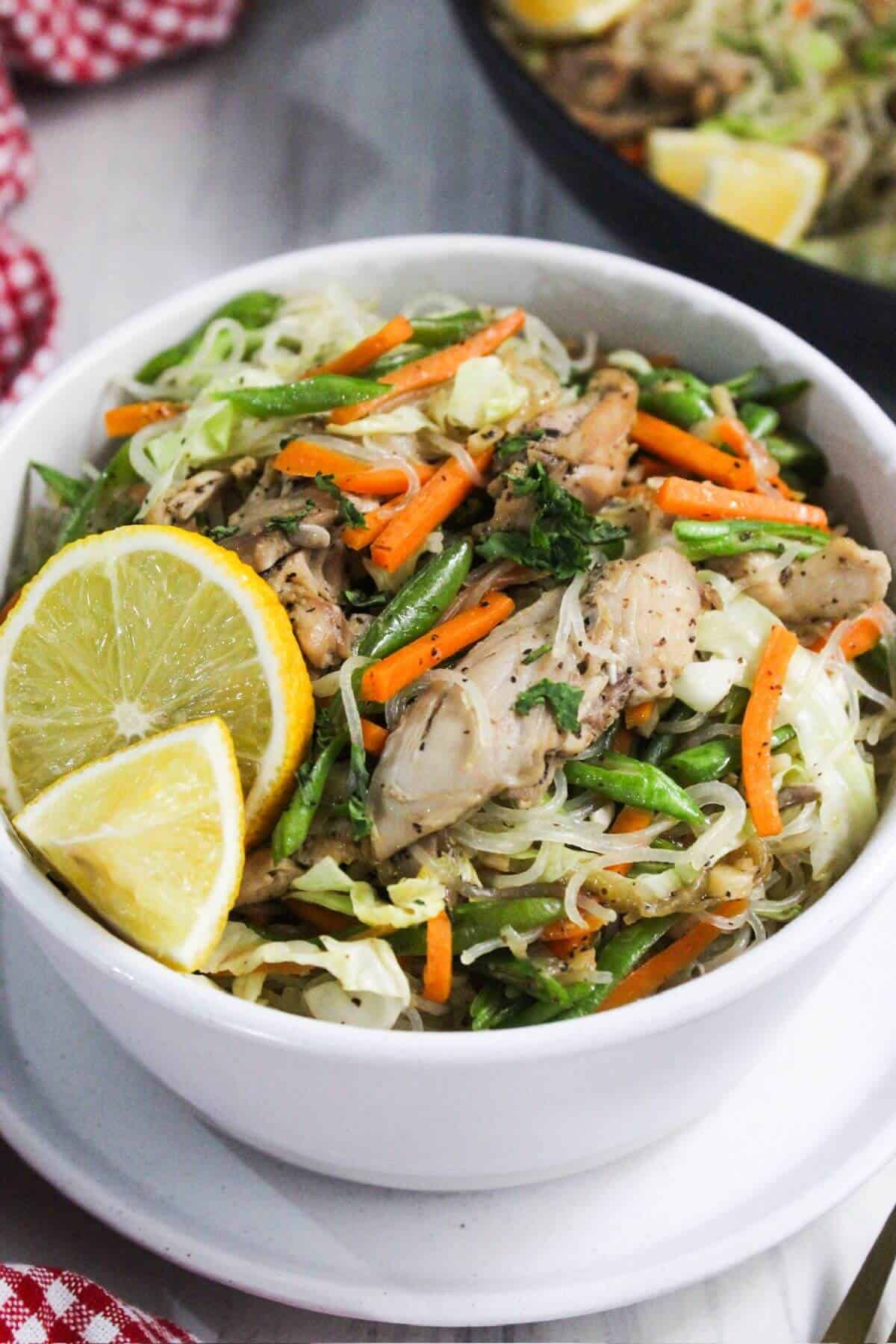 A white bowl with noodles, chicken, carrots, and lemon wedges.