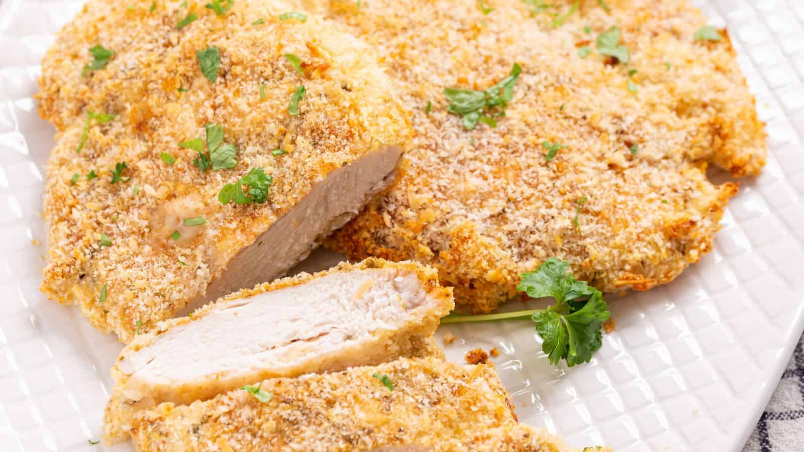 A close-up of a breaded, baked chicken breast cutlet garnished with parsley on a white plate.