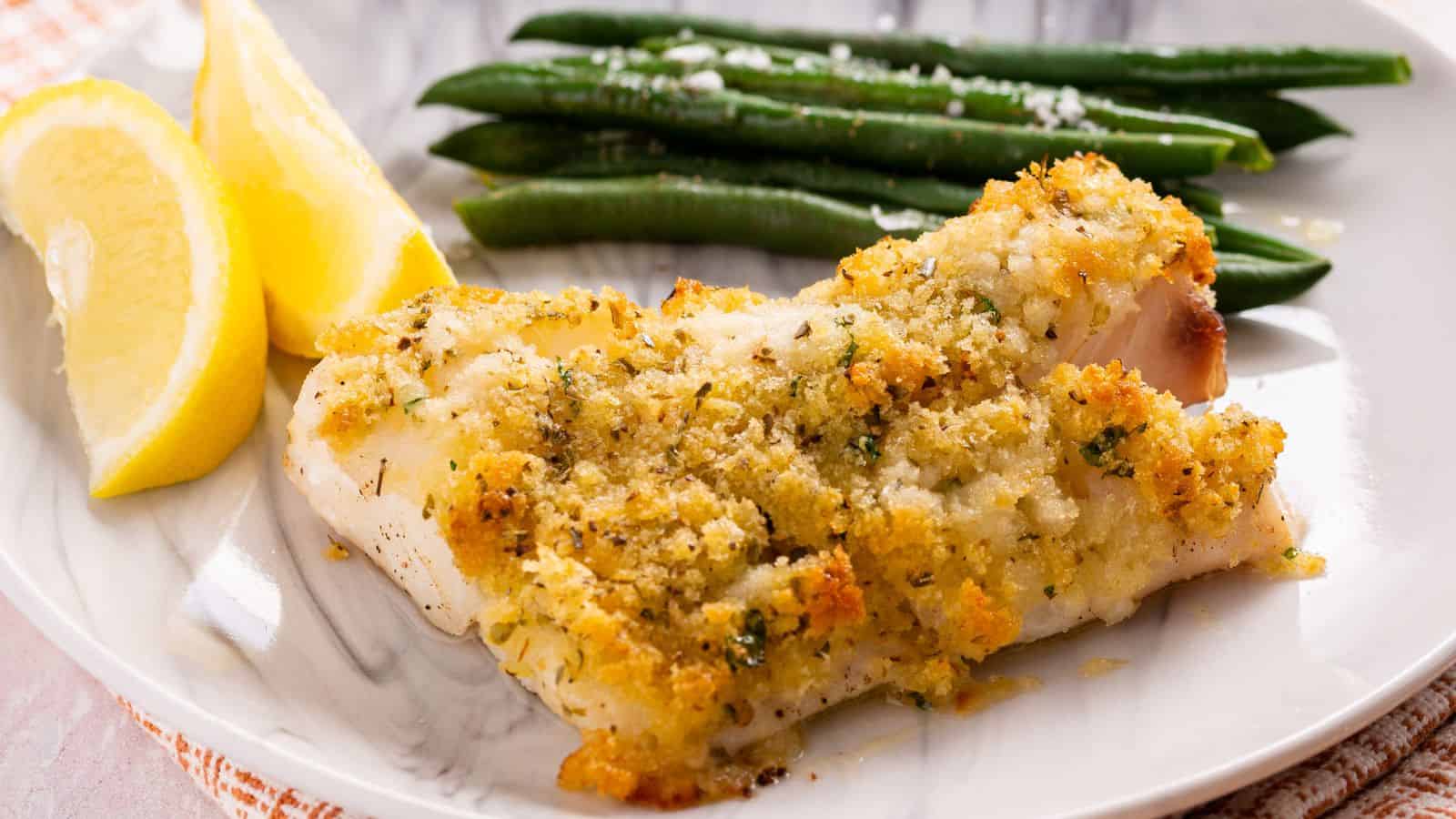 Breaded fish fillet served with green beans and lemon slices on a white plate.