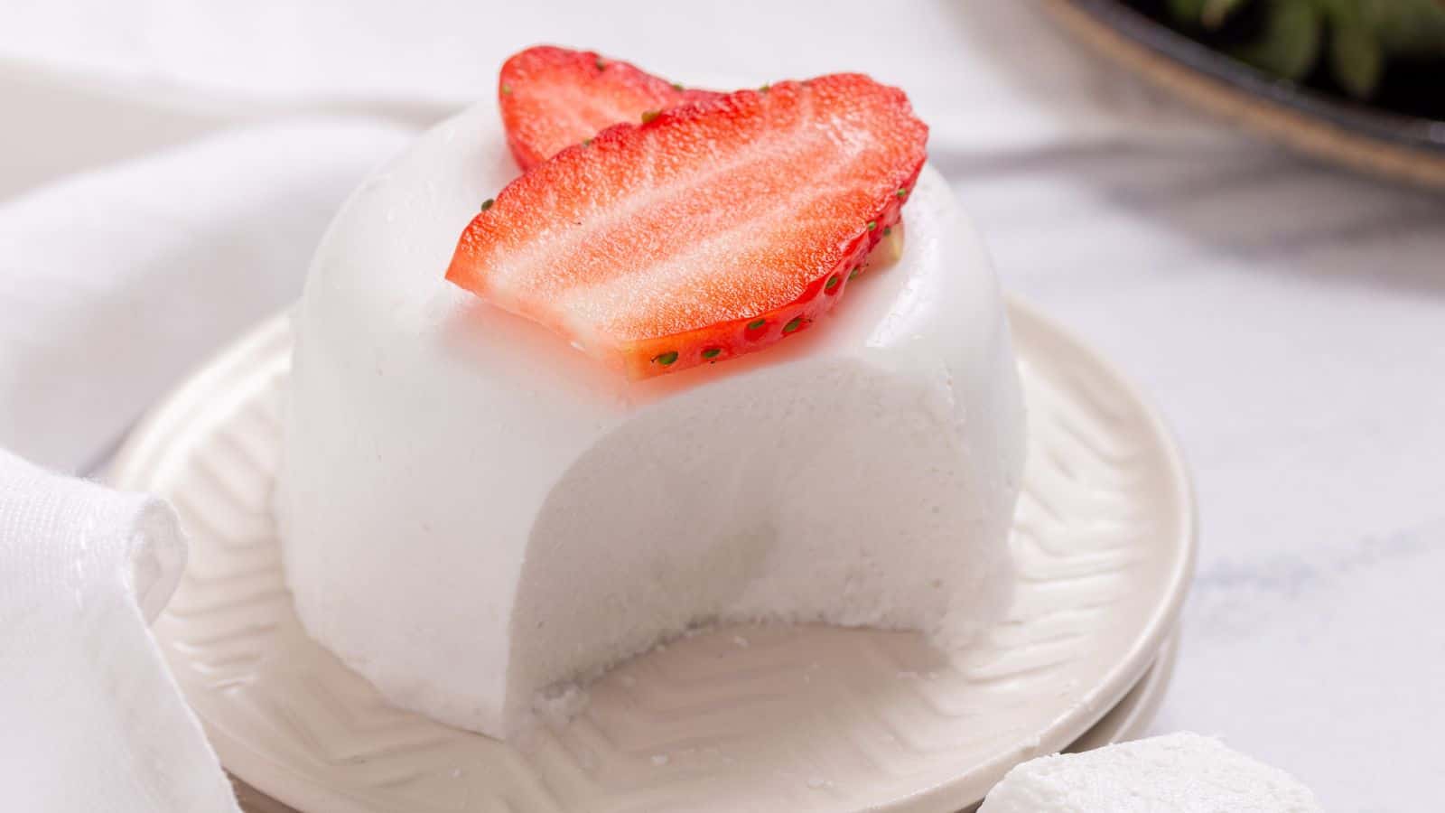 Panna cotta with spoonful taken out on plate topped with sliced strawberries.