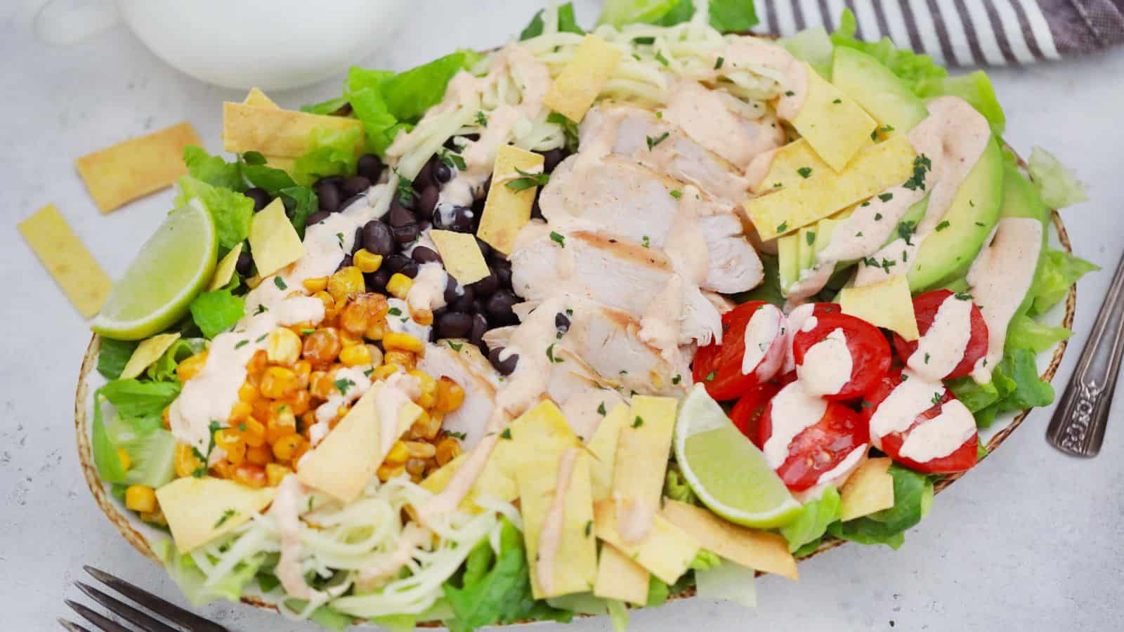A colorful chicken salad with avocado, black beans, corn, cherry tomatoes, and lime slices, presented in a white bowl.