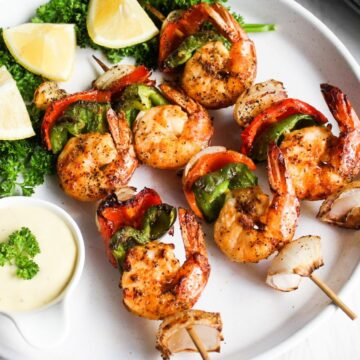 Grilled shrimp skewers with red and green bell peppers, served with lemon wedges and a dipping sauce on a white plate.