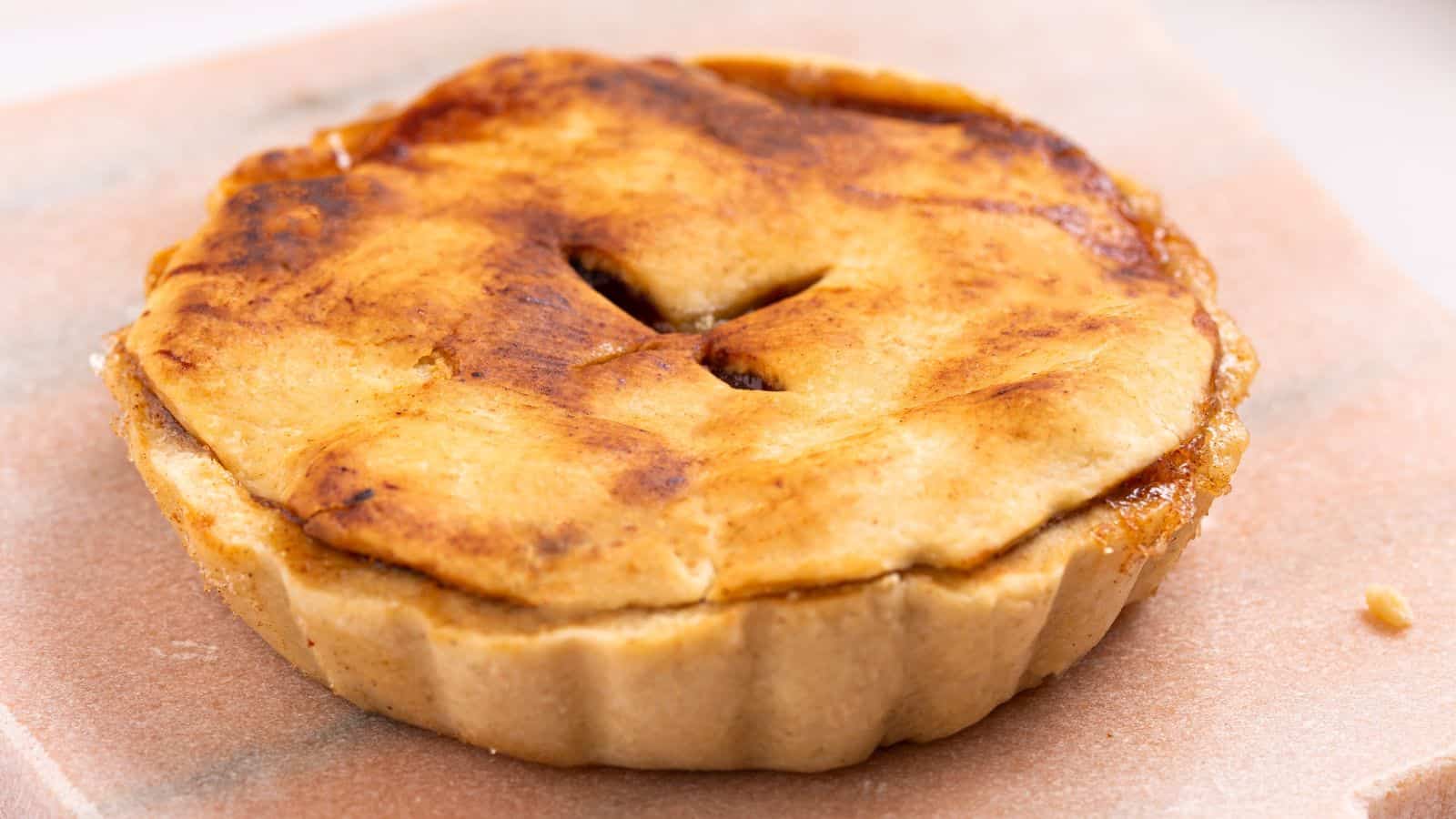 Golden-brown baked apple pie with a flaky crust and a cutout in the center, served on a beige surface.