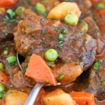 A close-up of a stew with beef chunks, carrots, peas, and potatoes, garnished with herbs.