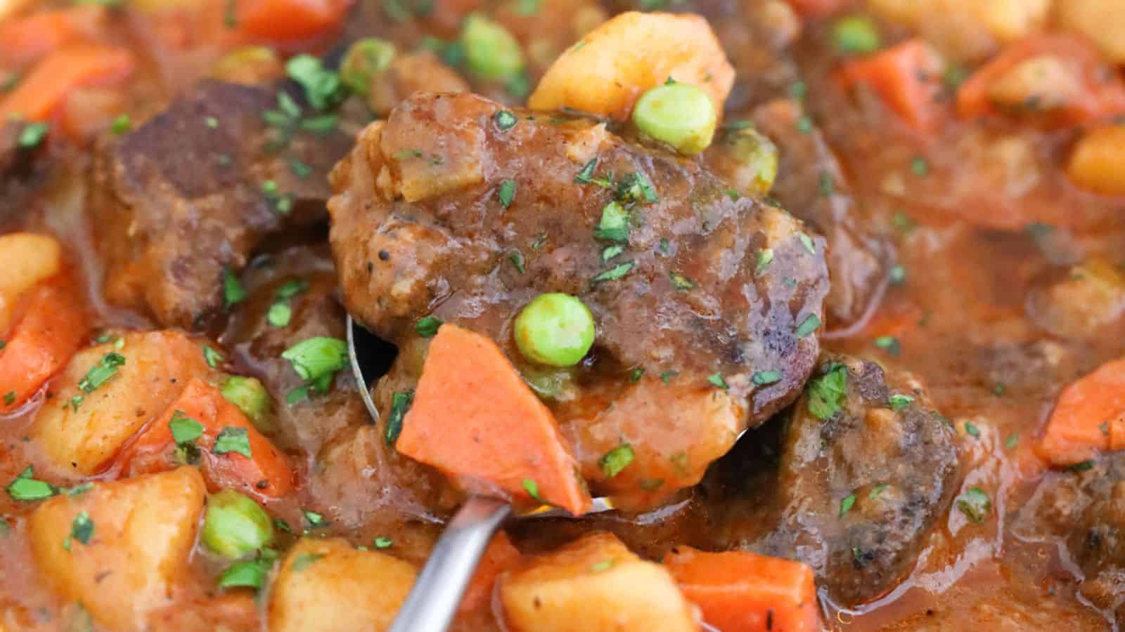 A close-up of a stew with beef chunks, carrots, peas, and potatoes, garnished with herbs.