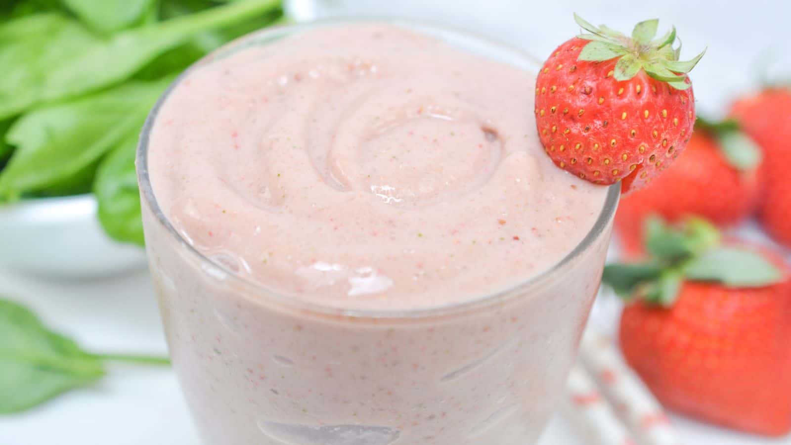 A glass of strawberry smoothie garnished with a whole strawberry, surrounded by fresh strawberries and spinach leaves.