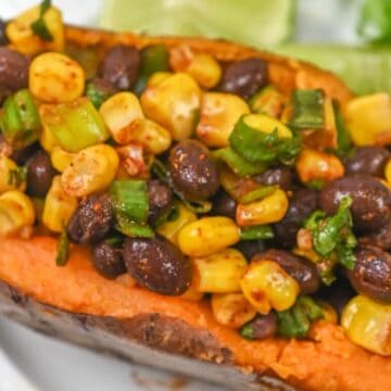 A roasted sweet potato topped with black beans, corn, and green onions, garnished with lime wedges on a white plate.