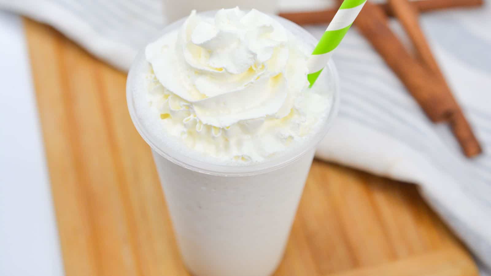 A whipped cream-topped milkshake with a striped straw, accompanied by cinnamon sticks on a wooden surface.