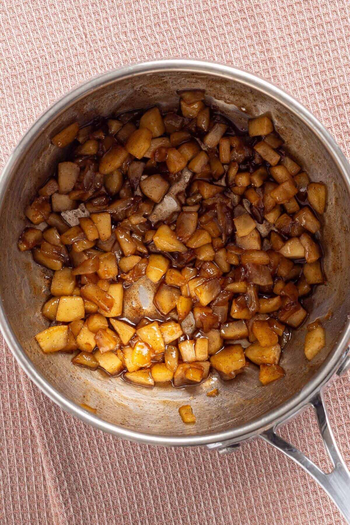 Cooked diced apples with cinnamon in a stainless steel pan on a textured tablecloth.