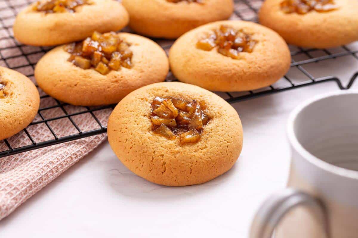 Freshly baked apple pie cookies topped with caramelized fruit on a cooling rack, with a cup and a cloth nearby on a light colored surface.