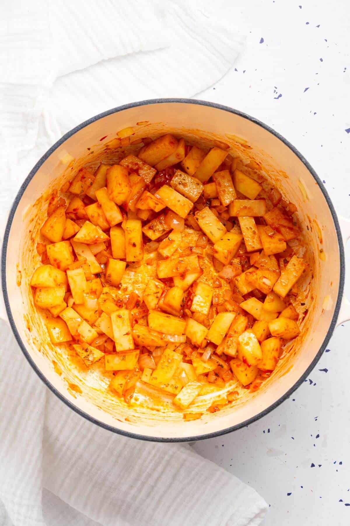 Potatoes mixed with tomato paste in a pot on a white surface.