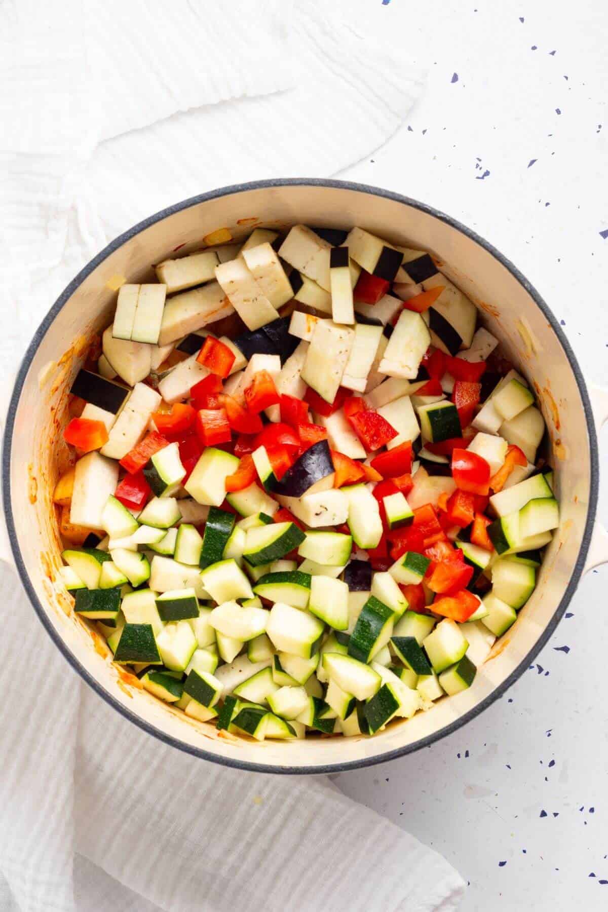 A pot containing chopped red peppers, zucchini, and eggplant on a white surface.