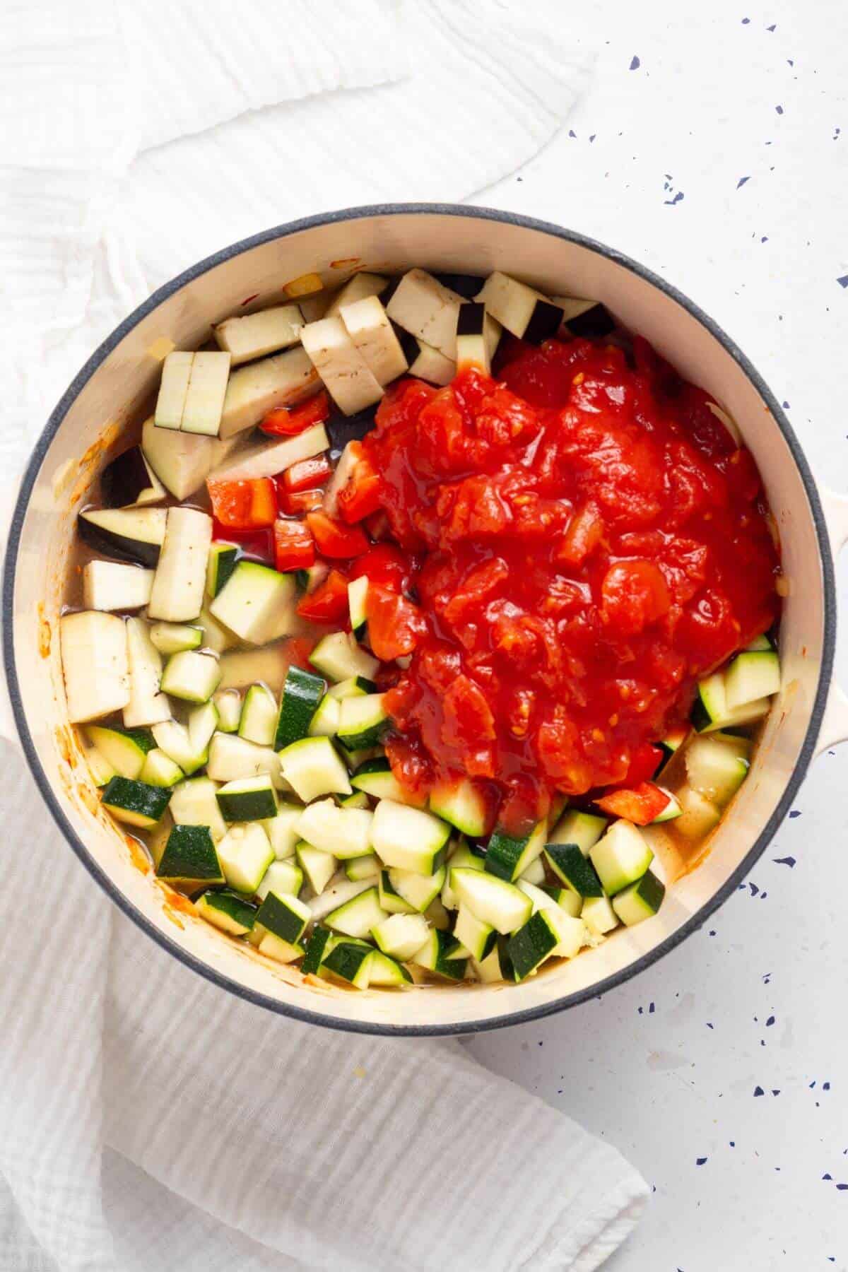 A pot with chopped zucchini and canned chopped tomatoes on a white surface, sprinkled with black pepper.