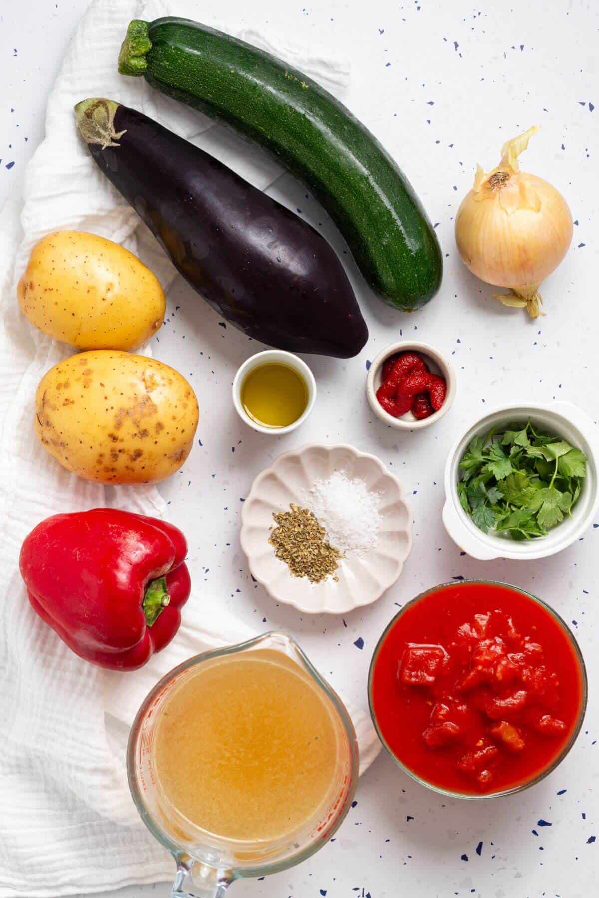 Top view of various fresh vegetables, including zucchinis, an eggplant, bell pepper, tomatoes, onions, and spices laid out on a white surface.