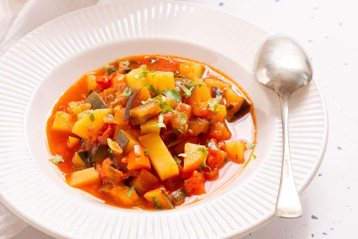 A bowl of ciambotta vegetable stew with diced vegetables and a garnish of herbs, served with a spoon on the side.