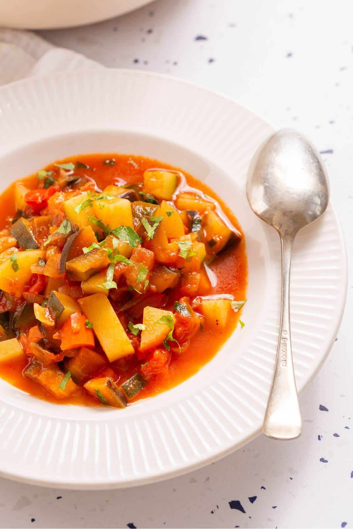 A plate of ciambotta vegetable stew with diced potatoes and red peppers, accompanied by a spoon, on a white speckled table.