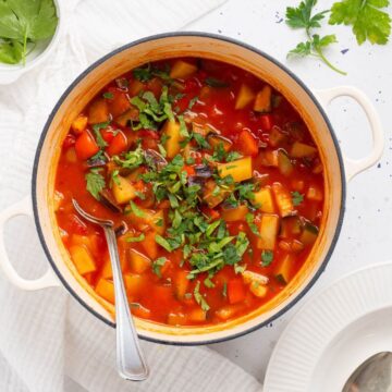 A vibrant ciambotta vegetable stew in a white pot, garnished with chopped herbs, with a spoon inside, served on a cloth-covered table.