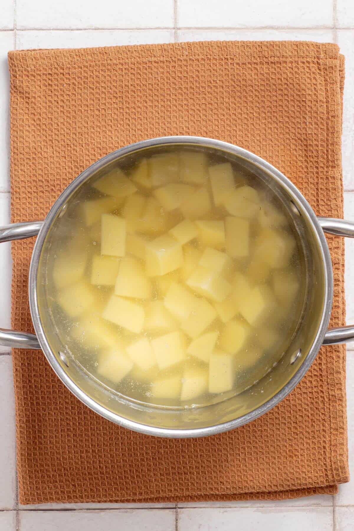 A stainless steel pot with diced potatoes in water, on an orange cloth atop a white tiled surface.