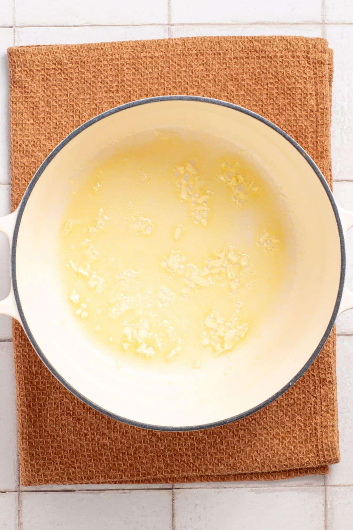Top view of a pot containing melted butter with garlic pieces, placed on a brown kitchen towel.