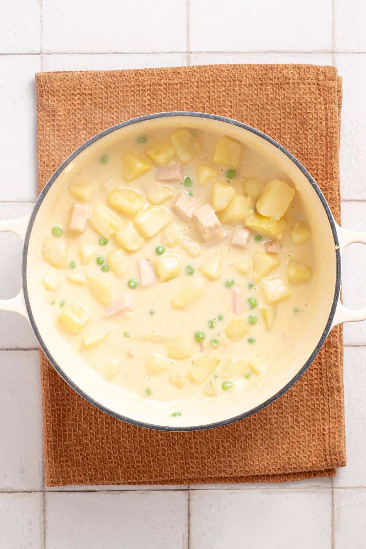 A pot of creamy ham and potato casserole with green peas on a brown cloth over a tiled surface.