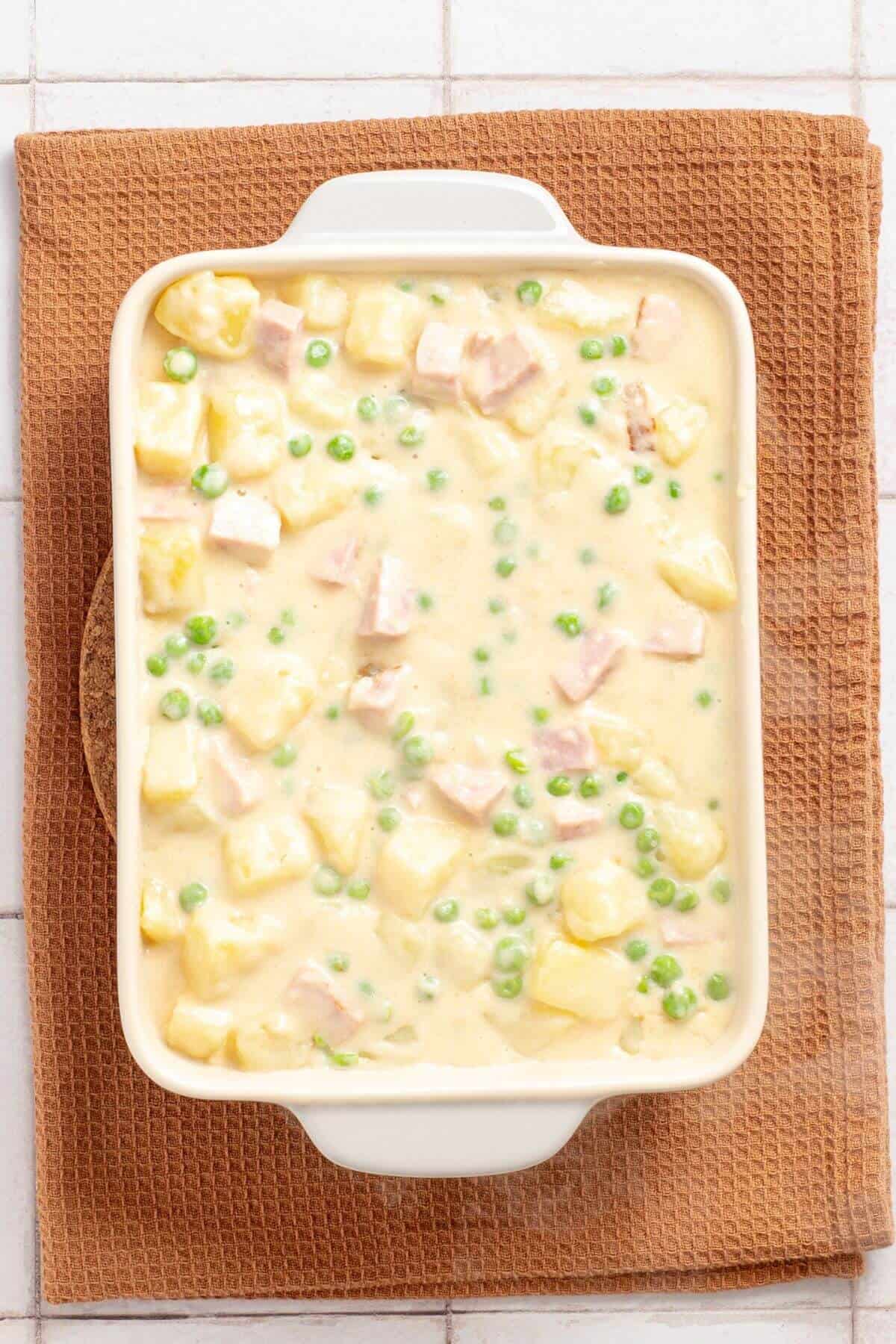 A casserole dish filled with creamy ham and potato casserole, presented on a brown cloth on a tiled countertop.