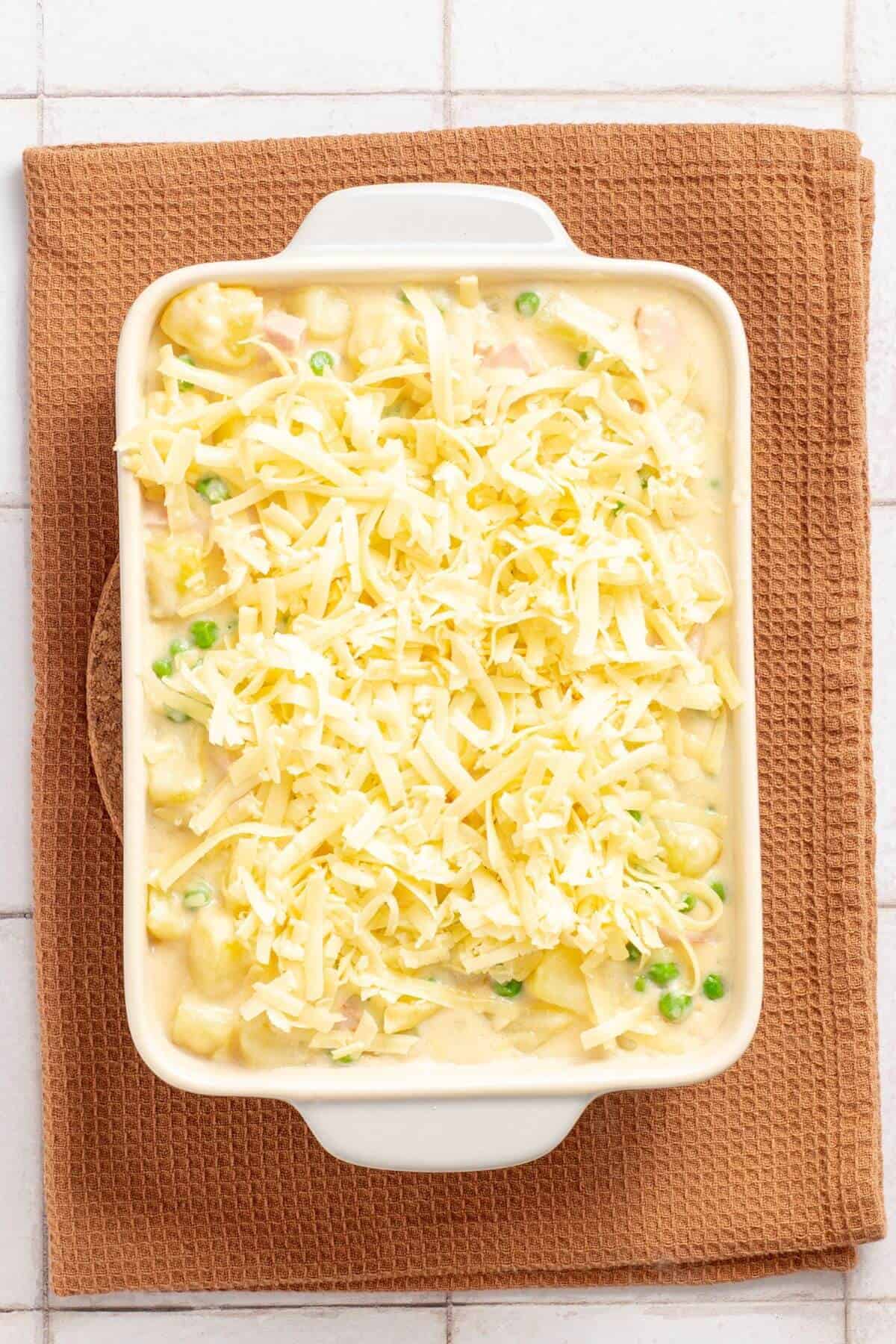 A casserole dish filled with a creamy ham, potato, and pea mixture, topped with shredded cheese, on a brown cloth.