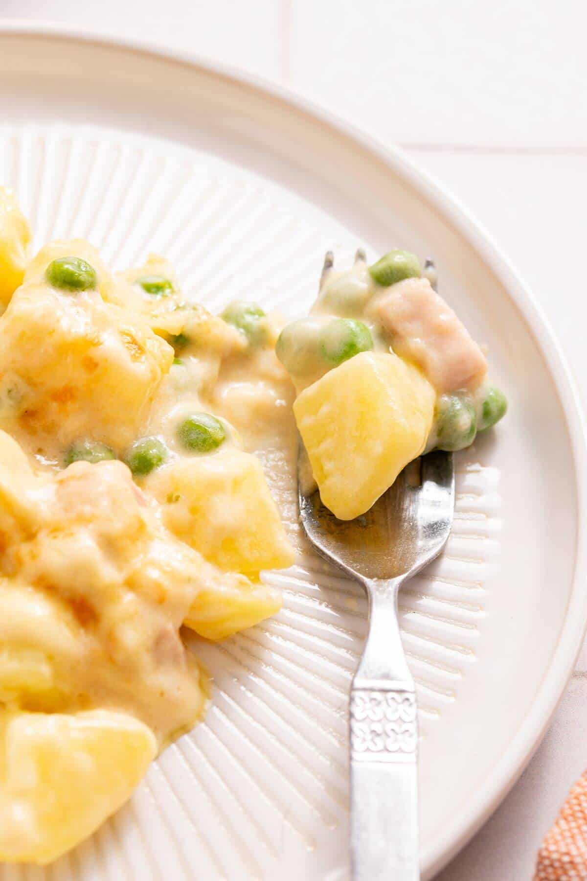 Creamy potato dish with peas and ham on a white plate with a fork.