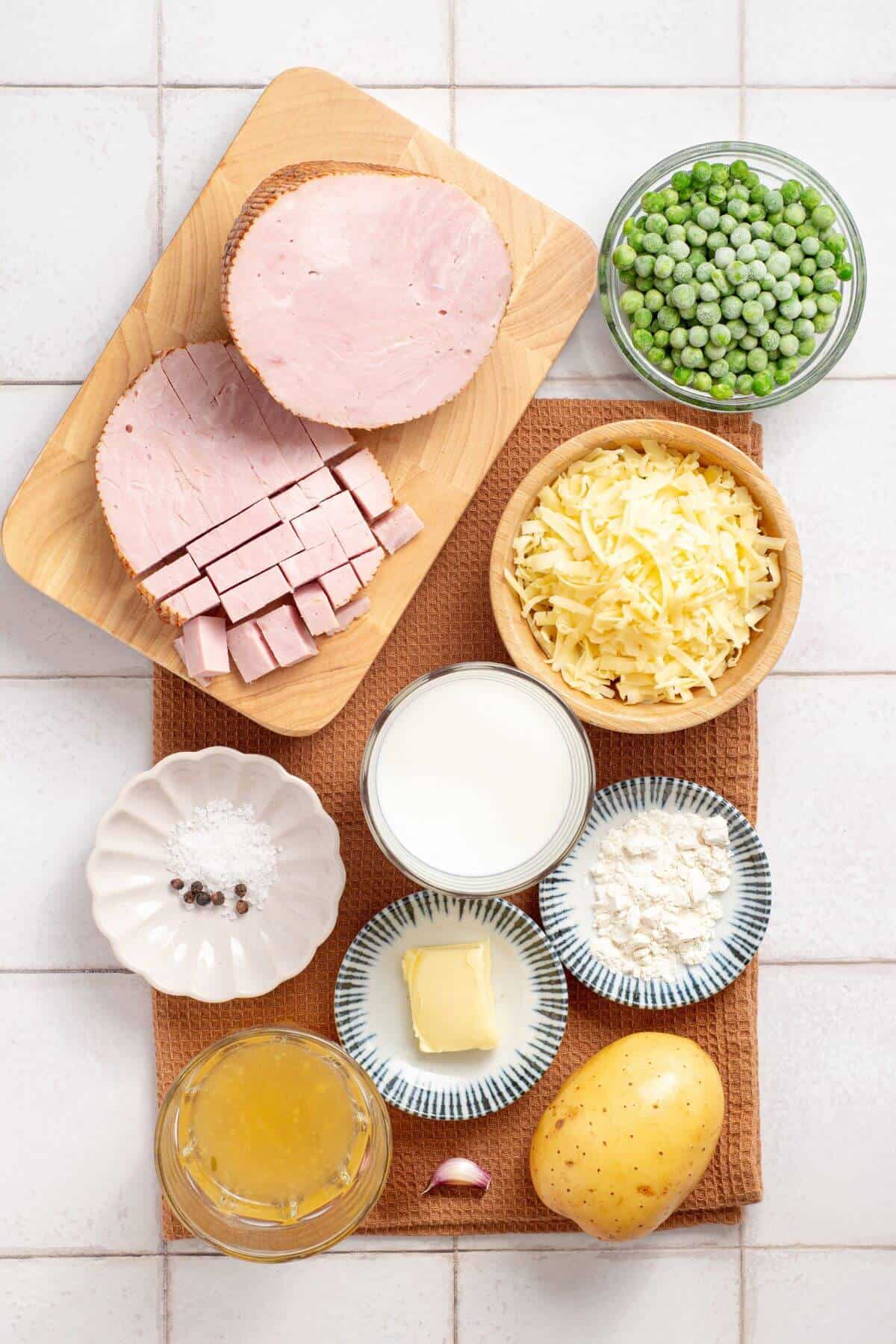 Various ingredients on a kitchen counter including sliced ham, peas, shredded cheese, broth, flour, butter, potatoes, and spices for making ham and potato casserole.