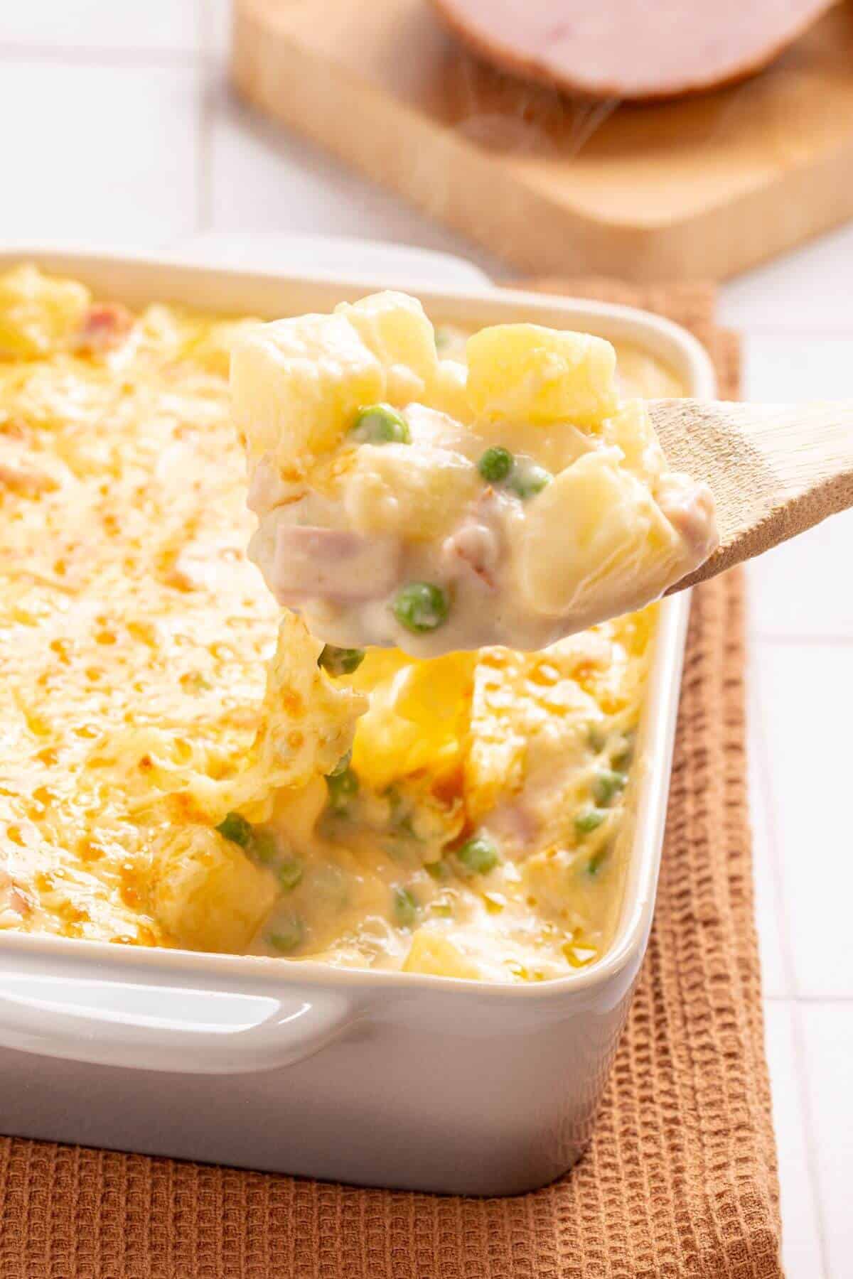 A spoon lifting a serving of creamy ham and potato casserole with corn, peas, and melted cheese from a white baking dish, with sliced ham in the background.