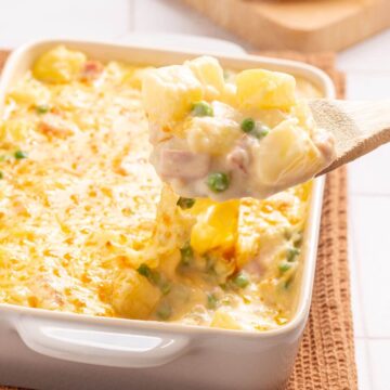 A baked ham and potato casserole with peas and cheese, served with a wooden spoon scooping a portion.