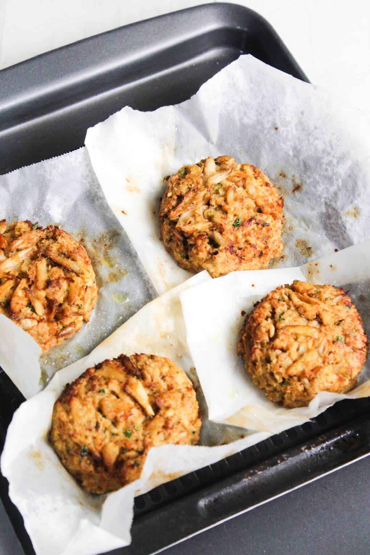 Four baked crab cakes on a baking tray lined with parchment paper.