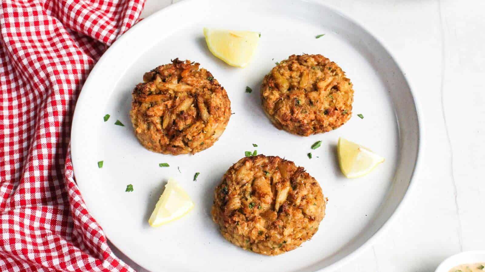 Four crab cakes on a white plate with lemon wedges, garnished with chives, on a red and white checkered cloth.