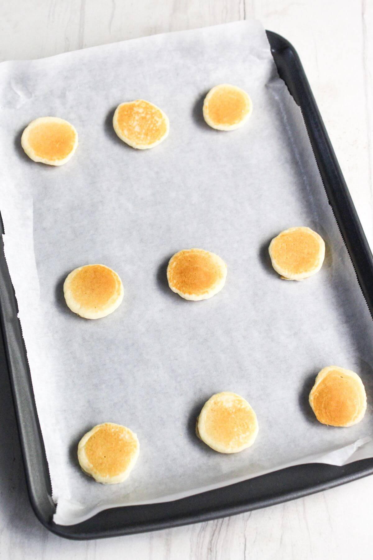 A baking tray lined with parchment paper holds nine mini pancakes spaced evenly.