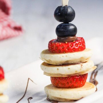 Three kebabs made of mini pancakes, strawberries, blueberries, and chocolate, arranged on a white plate with a red checkered cloth in the background.