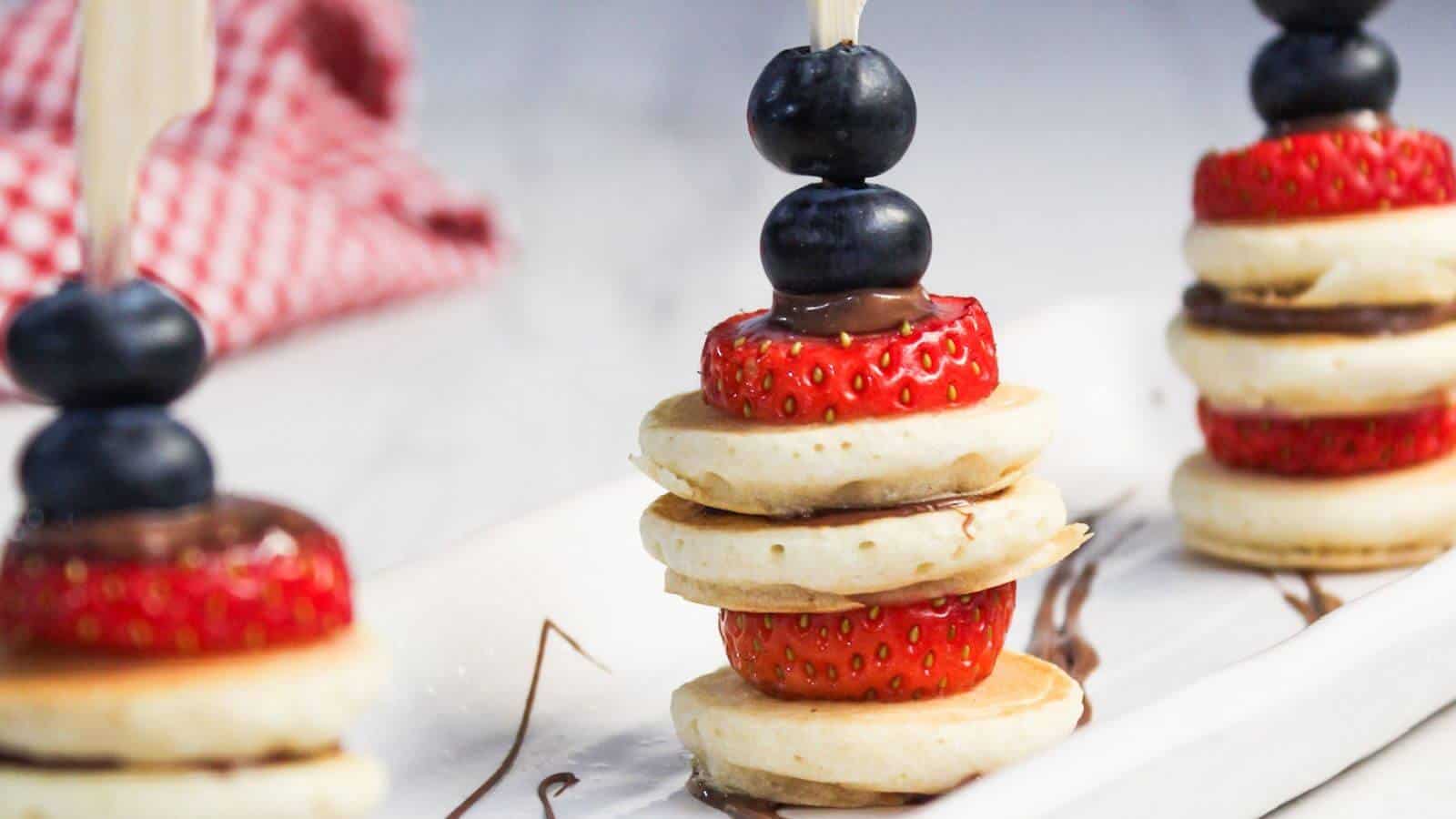 Three mini pancake stacks on skewers with layers of strawberries, blueberries, and chocolate spread, arranged on a white plate with a red checkered cloth in the background.