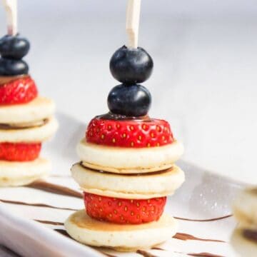 Small skewers holding bite-sized pancakes, strawberries, and blueberries arranged vertically on a white plate.