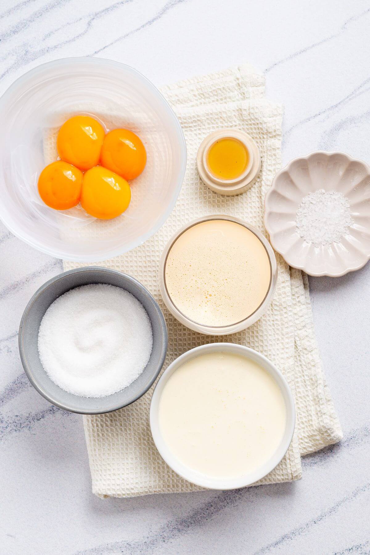 Overhead view of no churn ice cream ingredients, including egg yolks in a bowl, sugar, yeast, and milk, arranged neatly on a white marble surface.