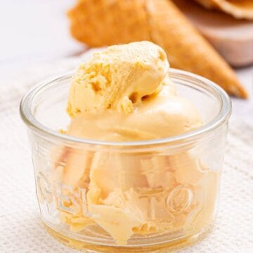 A scoop of mango no churn ice cream in a clear glass bowl, with a biscuit cone in the background on a white surface.