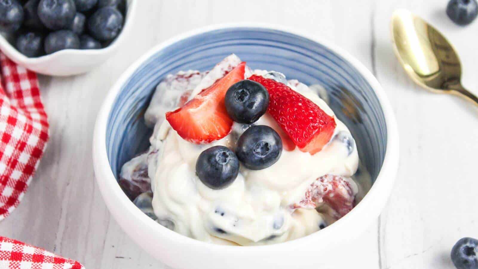 A bowl of creamy fruit salad with fresh strawberries and blueberries. A gold spoon and a small bowl of blueberries are on the side, along with a red and white checkered napkin.