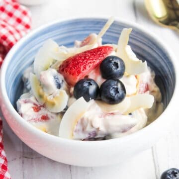 A bowl of creamy white salad topped with sliced strawberries, blueberries, and coconut flakes. A red and white checked cloth is visible to the left, along with a gold-colored spoon on the right.