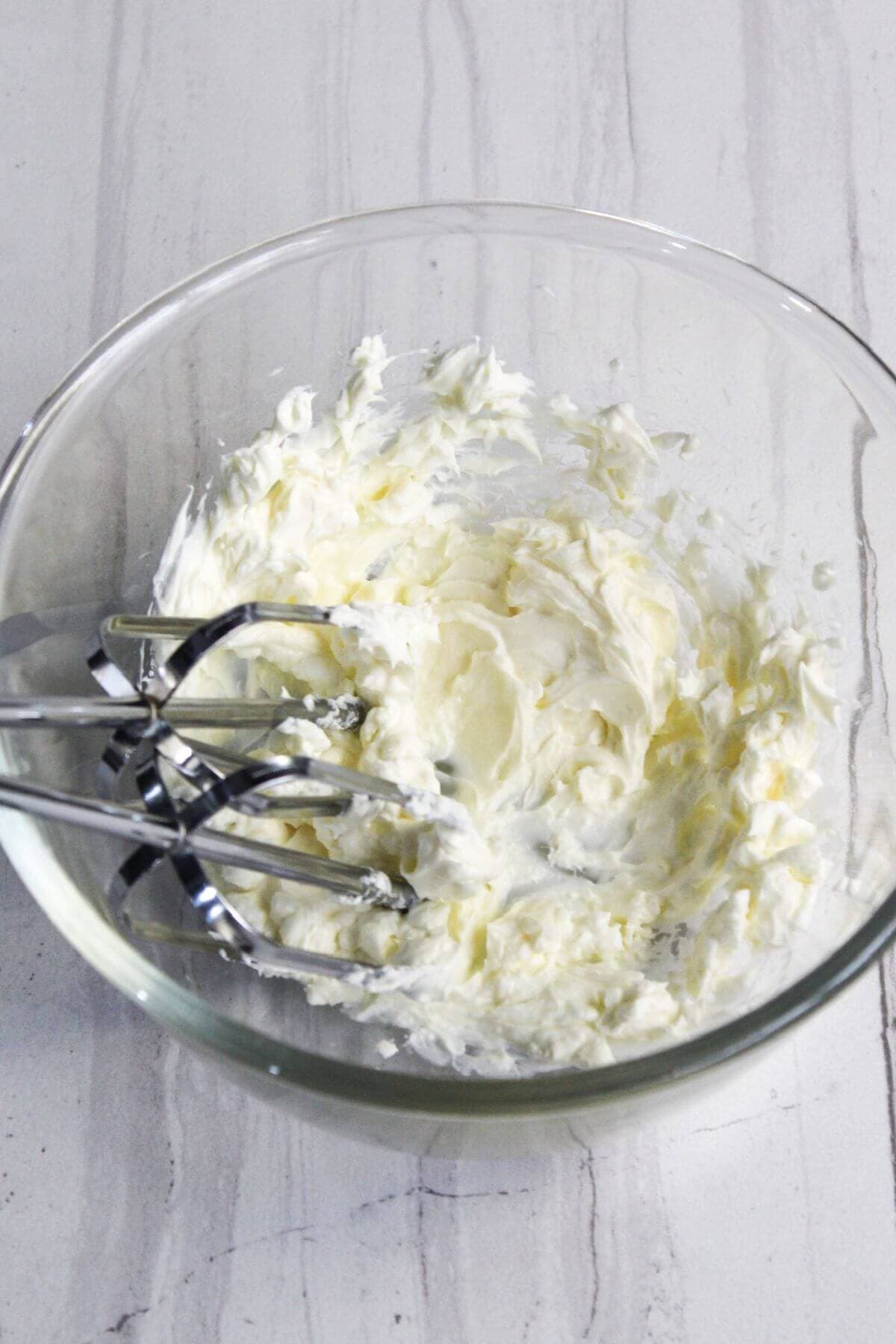 A glass bowl with whipped cream cheese and electric mixer beaters sitting inside it, placed on a white marble countertop.