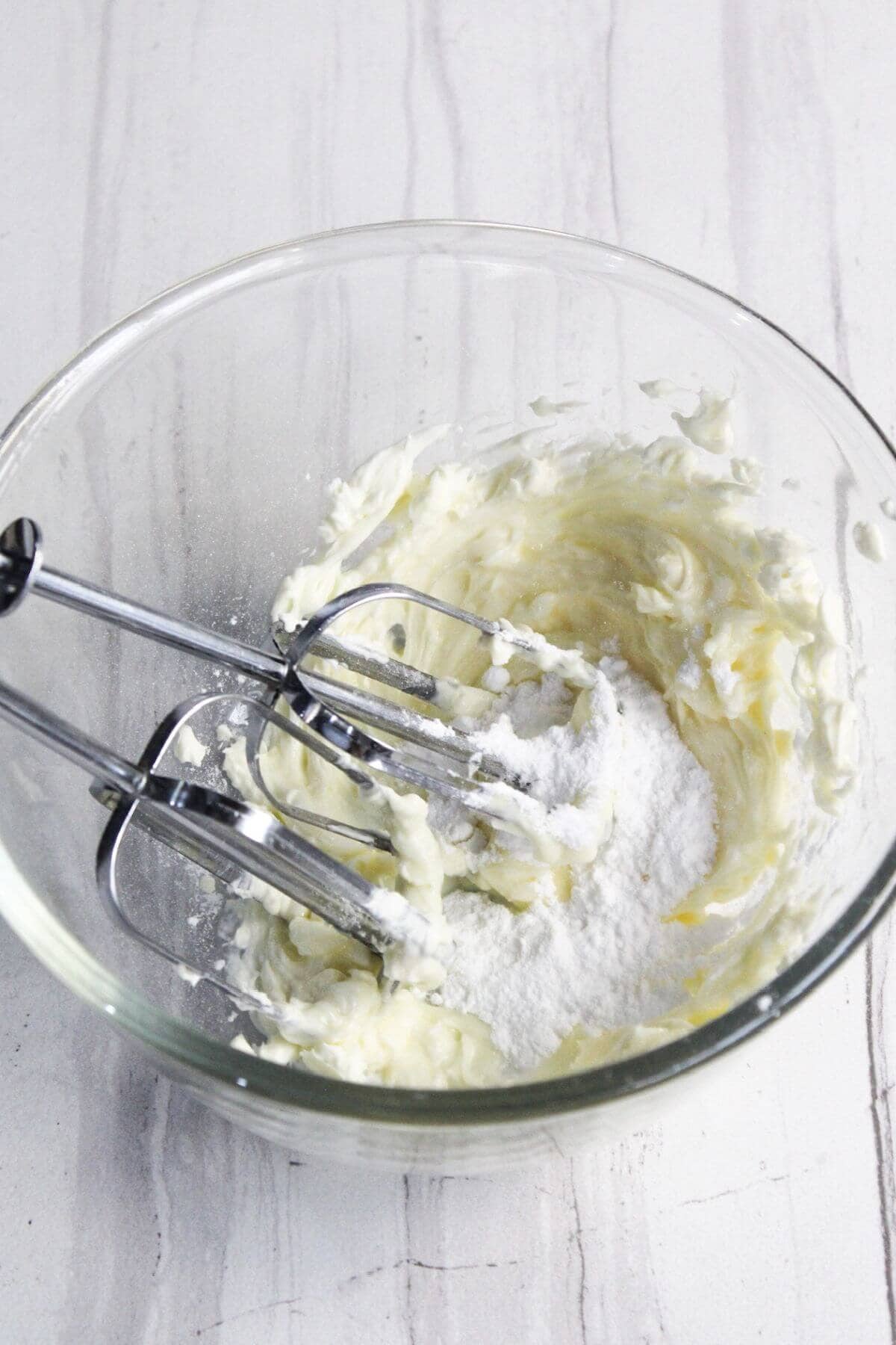 A glass mixing bowl with creamed butter and sugar, partially mixed, sits on a white surface. An electric hand mixer with beaters is placed in the bowl.