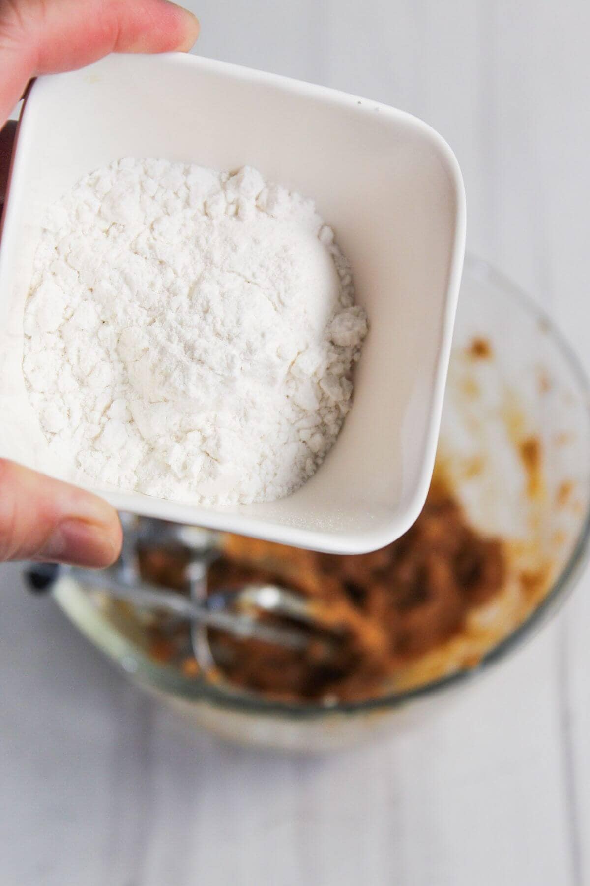 A person holds a white bowl of flour above a glass mixing bowl containing a brown mixture with a whisk attachment.